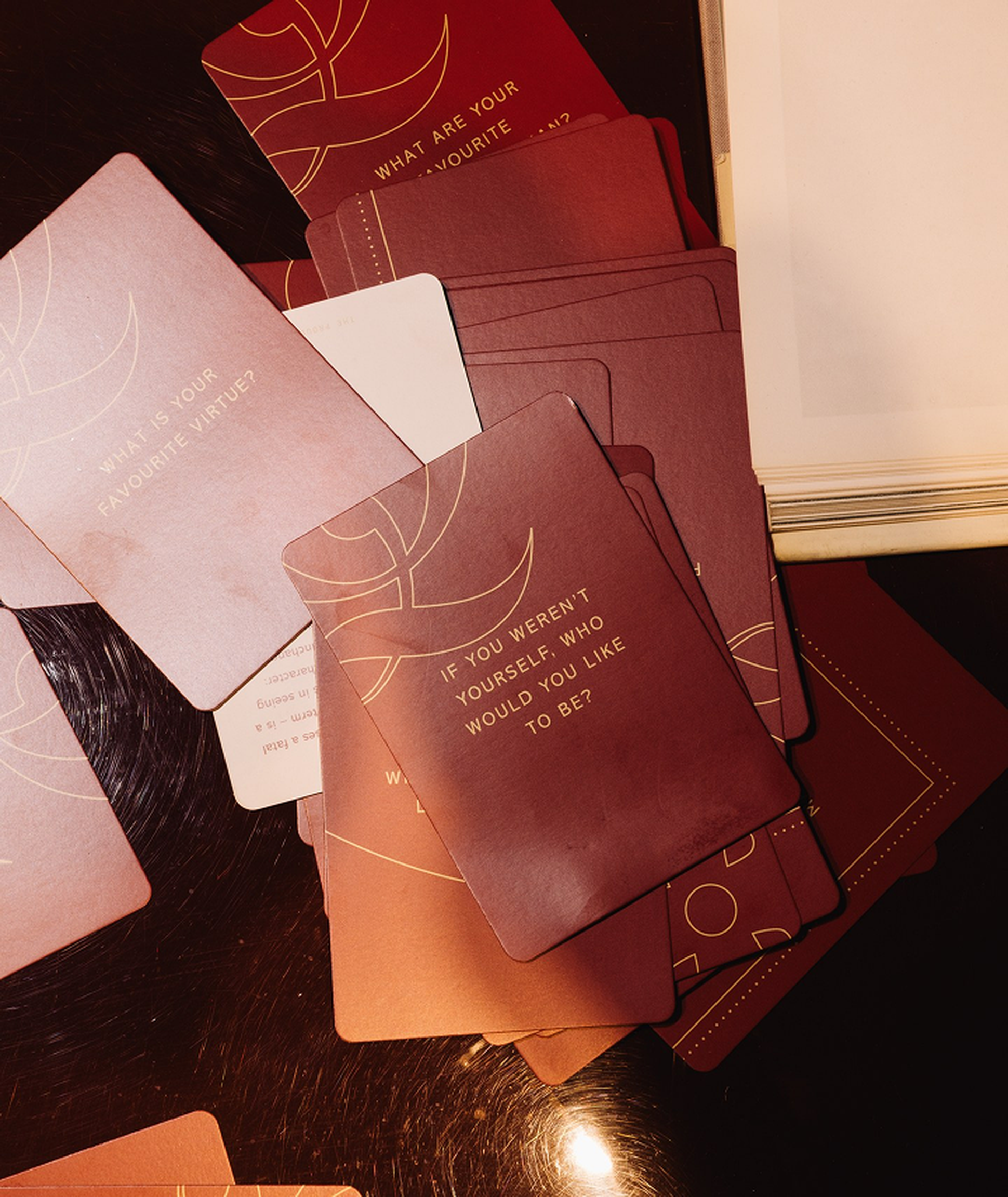 Scattered brown cards on a table, one prominently reading "IF YOU WEREN'T YOURSELF, WHO WOULD YOU LIKE TO BE?" in gold text.