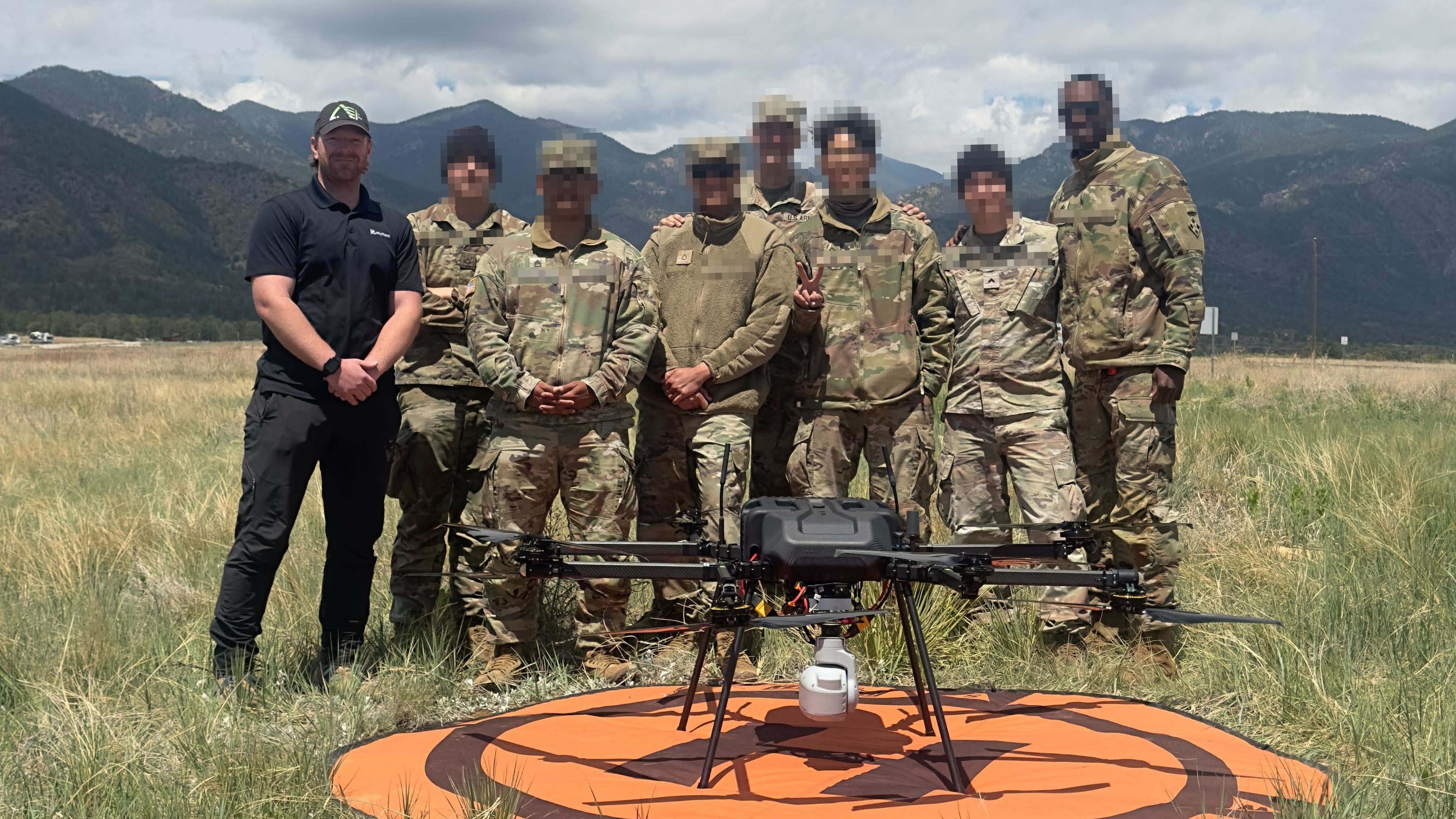 US Army soldiers stand on a grassy field with a hybrid drone, mountains in the background