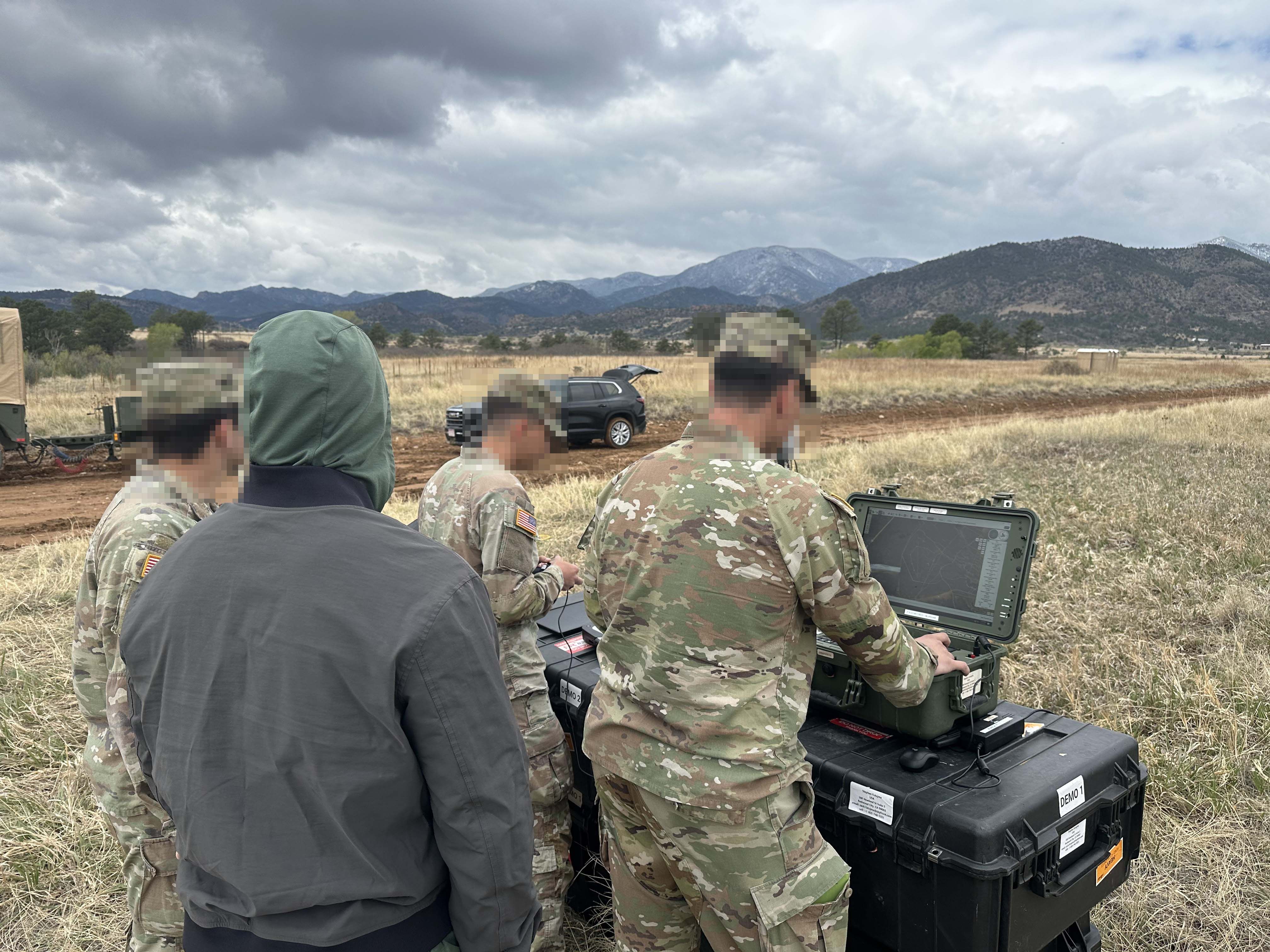 Military personnel hybrid drone GCS in a grassy field under cloudy skies, with mountains visible in the background.