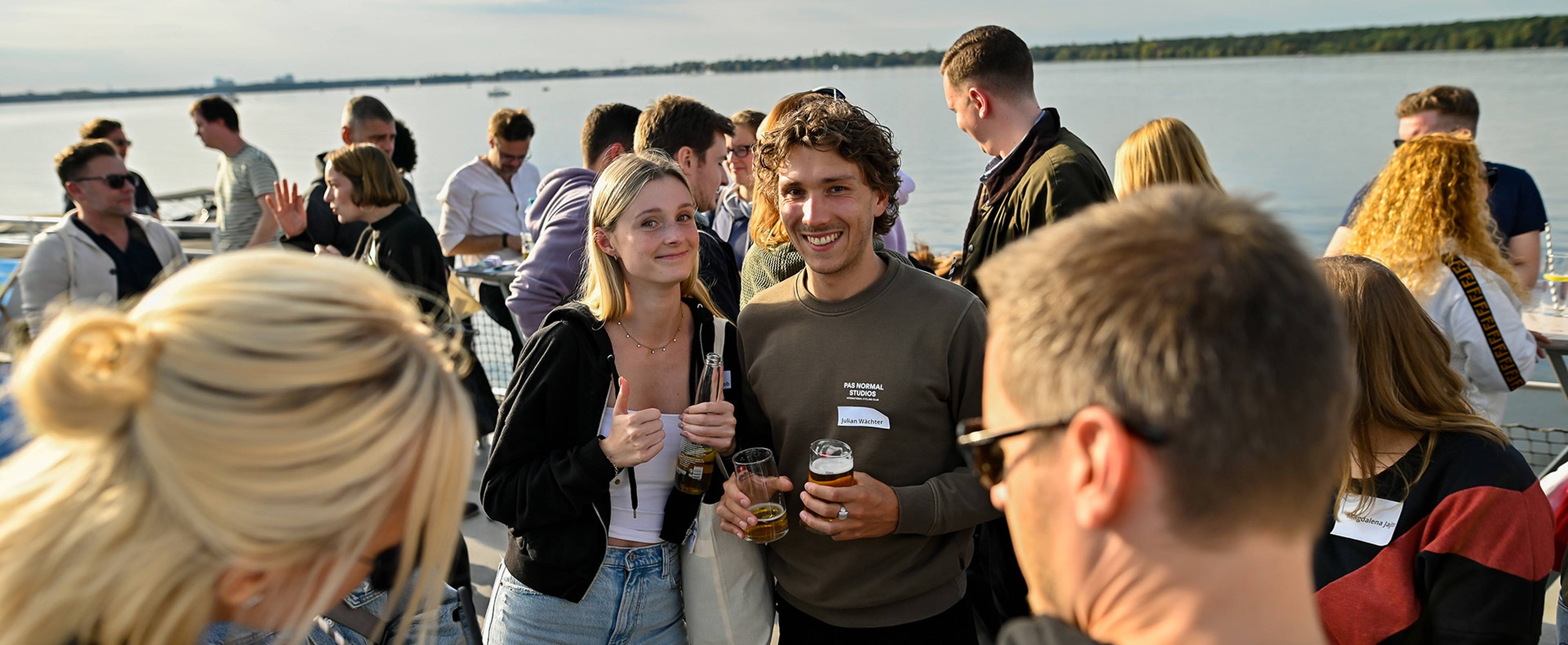 Maria und Julian lächeln mit Getränken auf einem Boot.