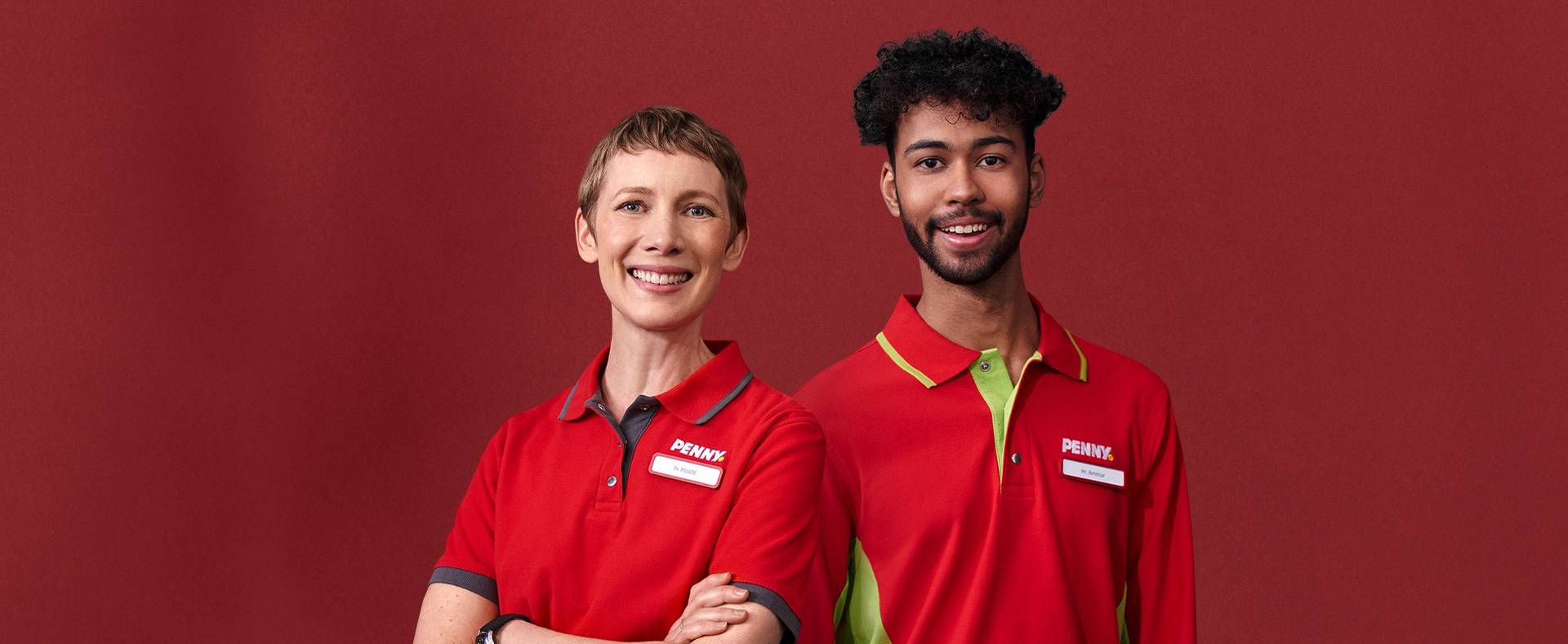 Zwei PENNY Mitarbeiter stehen vor einer roten Wand und lächeln in die Kamera. Sie tragen rote Arbeitskleidung mit dem PENNY Logo und einem Namensschild.