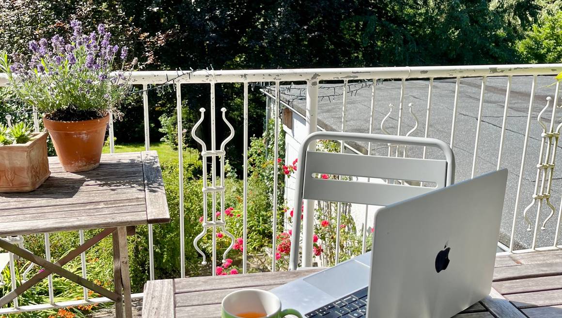 Laptop und Tasse auf Balkon, dahinter Pflanzen und großer Baum.