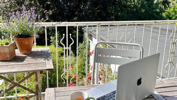 Laptop und Tasse auf Balkon, dahinter Pflanzen und großer Baum.