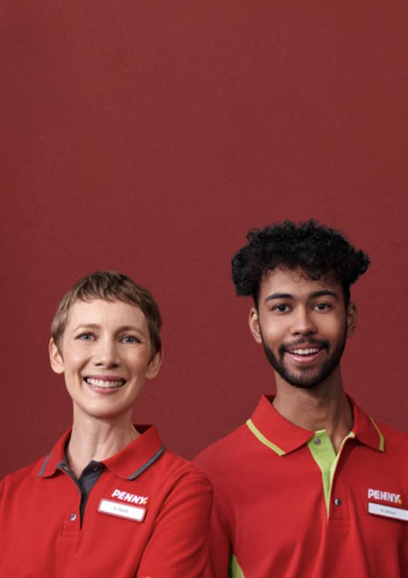 Zwei PENNY-Mitarbeitende in roter Arbeitskleidung mit Logo und Namensschild lächeln vor einer roten Wand.