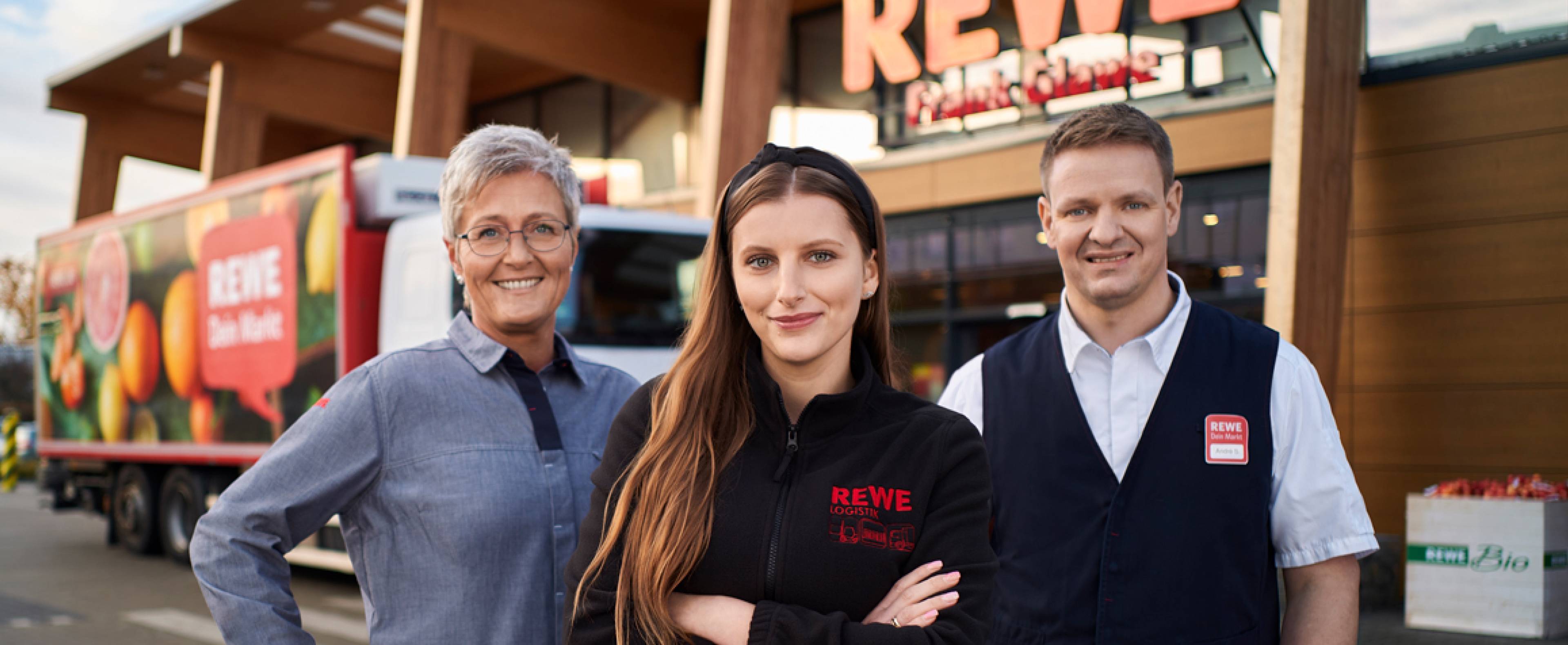 Eine Angestellte und ein Angestellter in REWE-Arbeitskleidung stehen nebeneinander und lächeln freundlich in die Kamera.