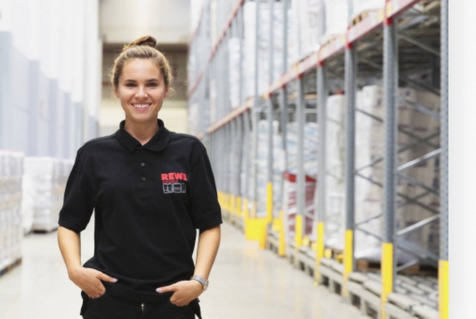Frau in schwarzer Kleidung mit rotem REWE-Logo lächelt in einer Lagerhalle.