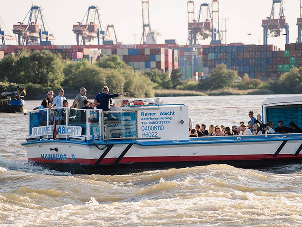 Blau-weißes Boot namens Rainer fährt auf der Elbe, mit Menschen an Bord und dem Hamburger Hafen im Hintergrund.