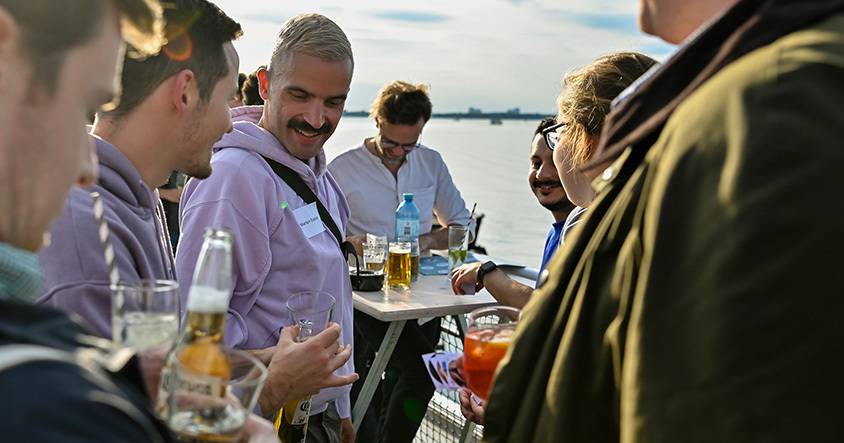 Marlon und Sebastian stehen zwischen mehreren Personen an Stehtischen, lachen und halten Getränke in der Hand.