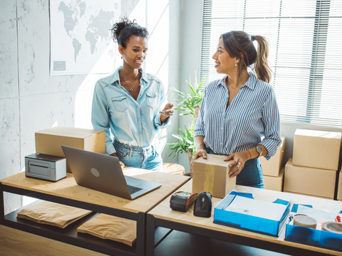 Two women in a bright office packing boxes, with a laptop and shipping supplies on the desk. Sunlight streams through the window blinds.