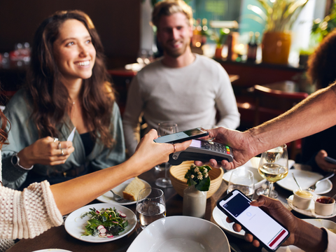 People at a restaurant table, one person paying with a smartphone via contactless payment. Plates of food and drinks are on the table.