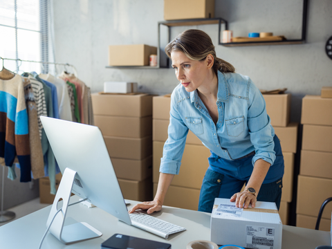 Woman in a denim shirt working at a computer in a room with boxes and clothes, holding a package.
