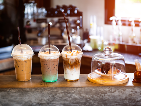 Three iced coffee drinks with straws on a counter next to a glass dome covering chocolate cake slices, in a cozy café setting.