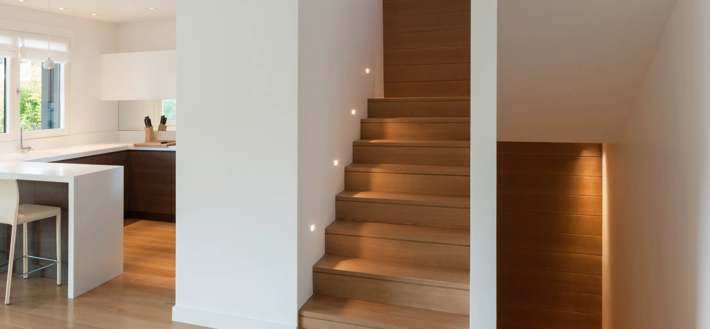 Moderne Holztreppe mit dezenten, quadratischen LED-Wandspots zur Stufenbeleuchtung neben einer offenen Küche
