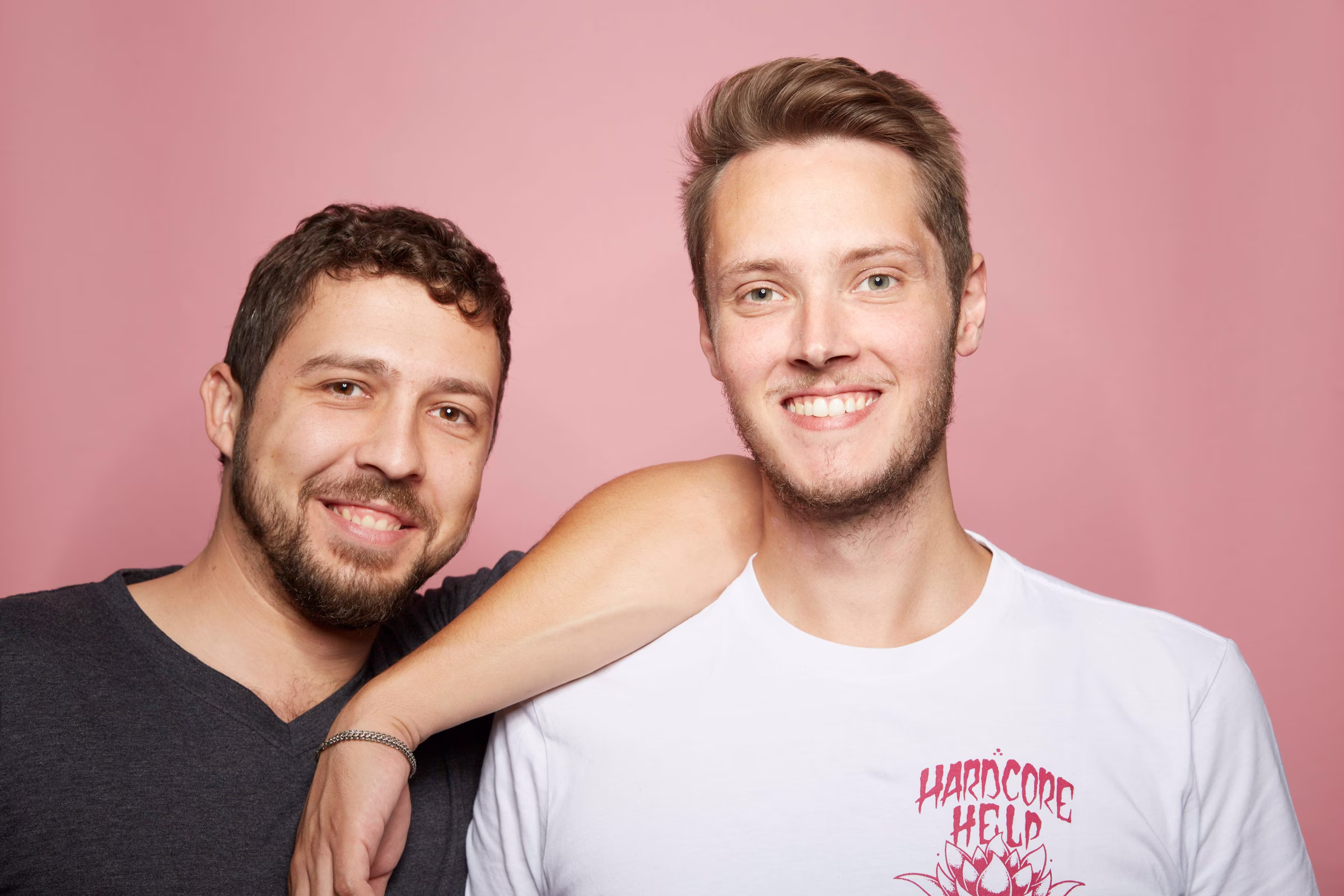 Two smiling men standing close together against a pink background, one wearing a dark shirt, the other in a white shirt with red lettering.