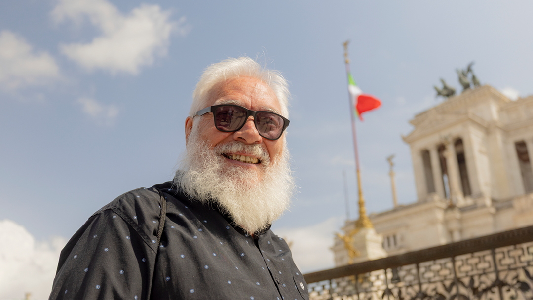 man with a white beard and sunglasses, standing outside in front of the Altare della Patria monument with an Italian flag.