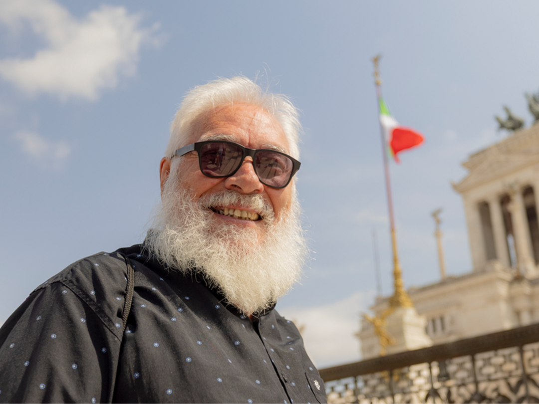 An older man with a white beard and sunglasses, standing outside in front of the Altare della Patria monument with an Italian flag.