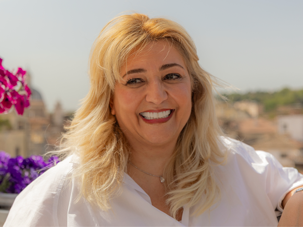 Mujer con cabello rubio sonriendo en una terraza soleada, con flores rosadas y una vista de la ciudad visible al fondo.
