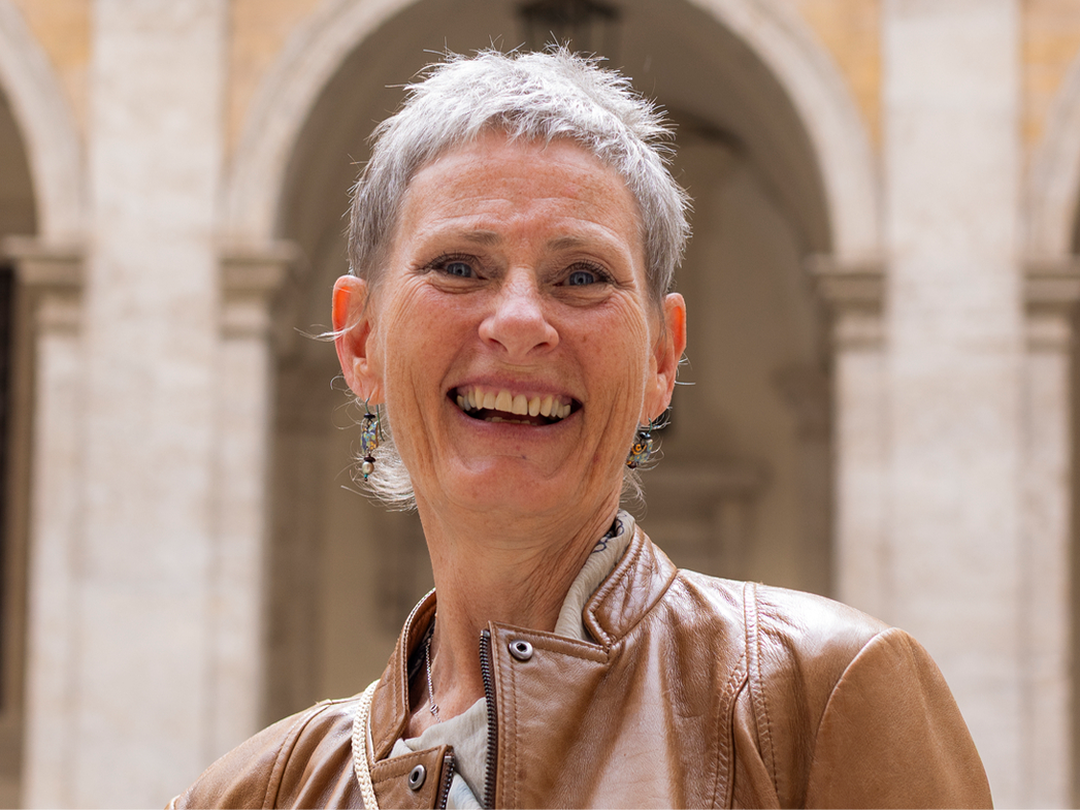 A woman with short grey hair smiling, with a classical Italian courtyard with arched colonnades visible behind her.