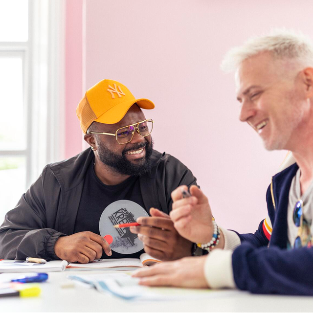 Two men sitting at a table together, one wearing an orange cap, both smiling during what appears to be a class activity.