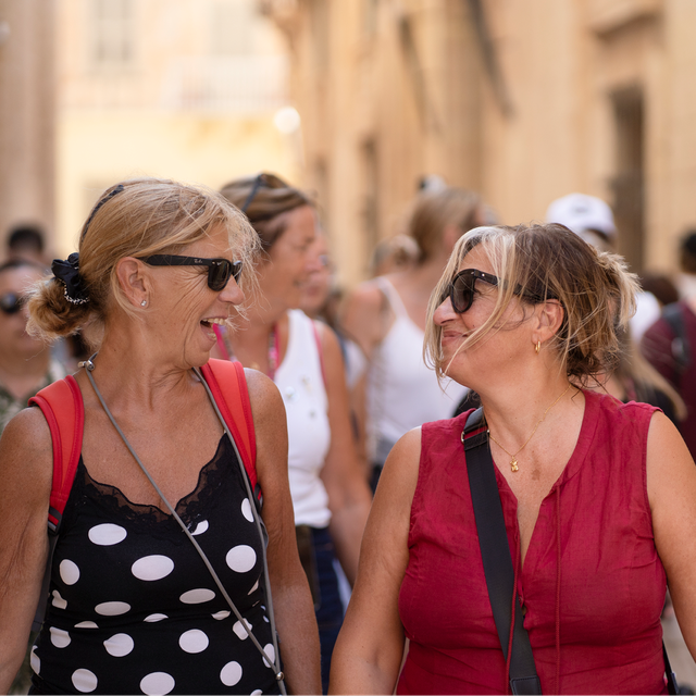 Two women wearing sunglasses walking together through a busy narrow street lined with stone buildings.