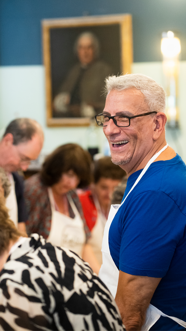 A man with white hair and glasses wearing an apron, smiling in a room with other people and a portrait painting on the wall.