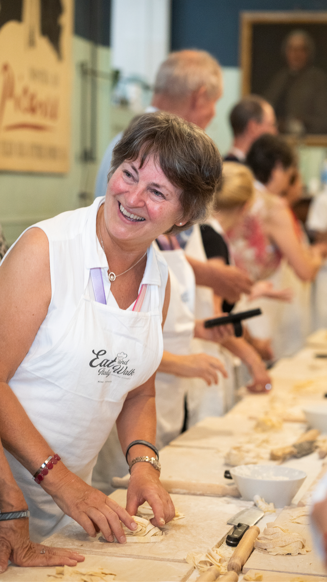 A woman in a white apron making pasta at a long table during a cooking class with other participants.