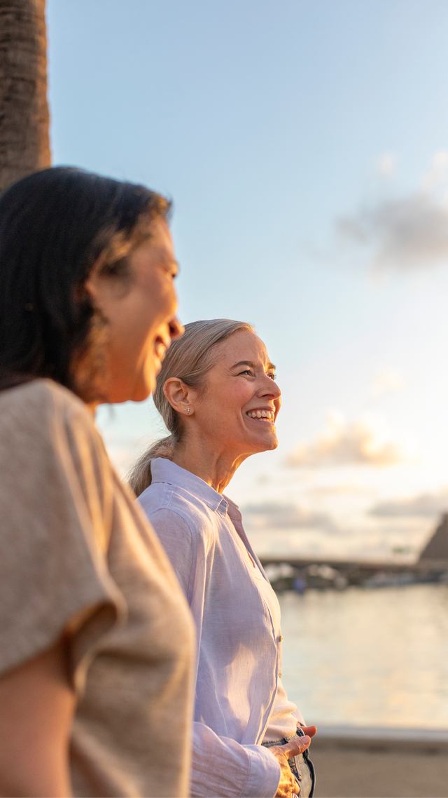 Two women standing near a waterfront at sunset, looking into the distance with a palm tree beside them.