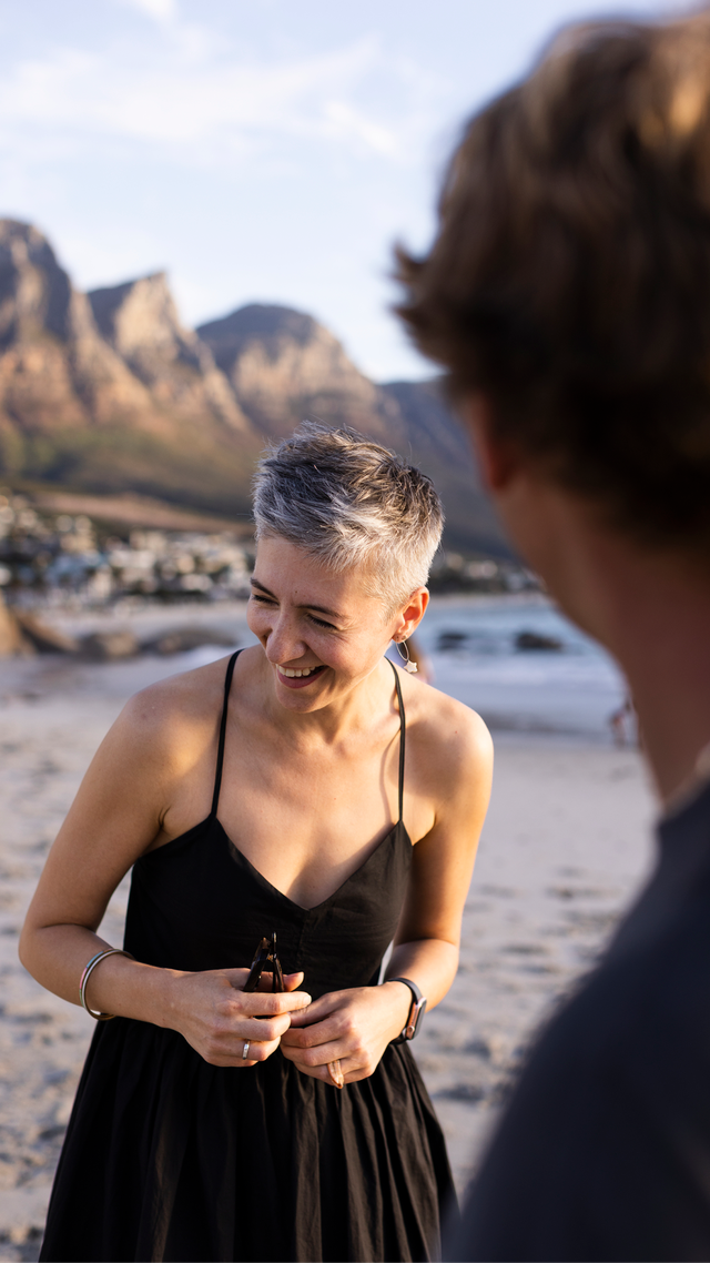 A woman with short grey hair smiling, standing on a beach facing another person, with mountains visible in the background.