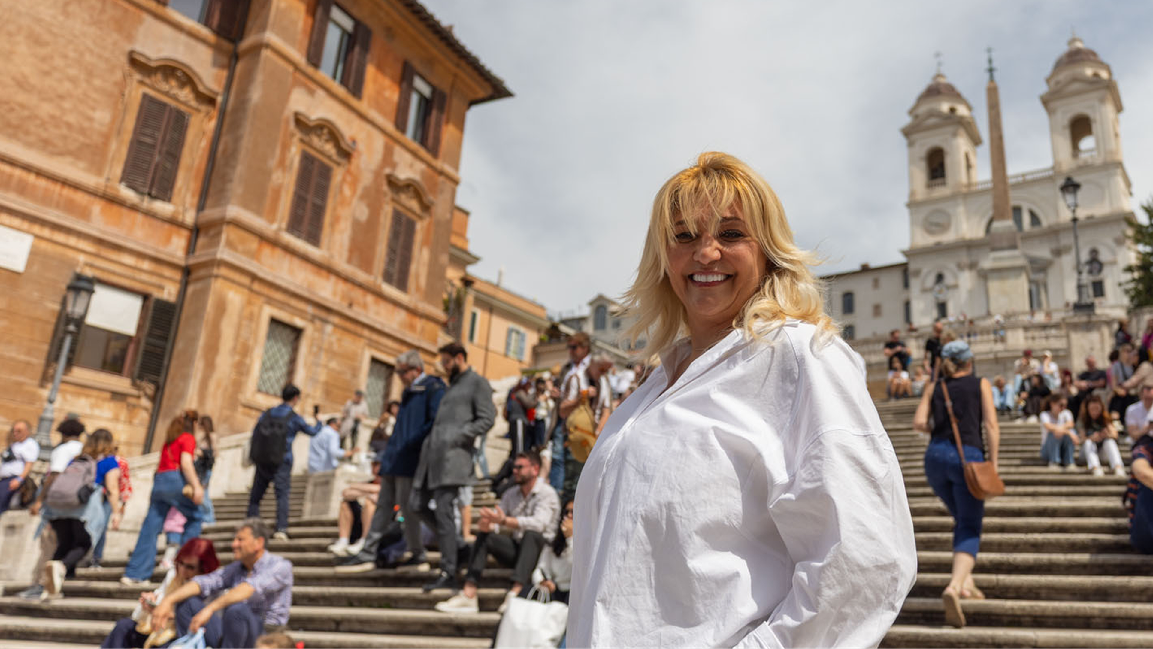 A woman with blonde hair and sunglasses in a white shirt standing on the Spanish Steps in Rome, with a crowd of people and the Trinità dei Monti church visible behind her.
