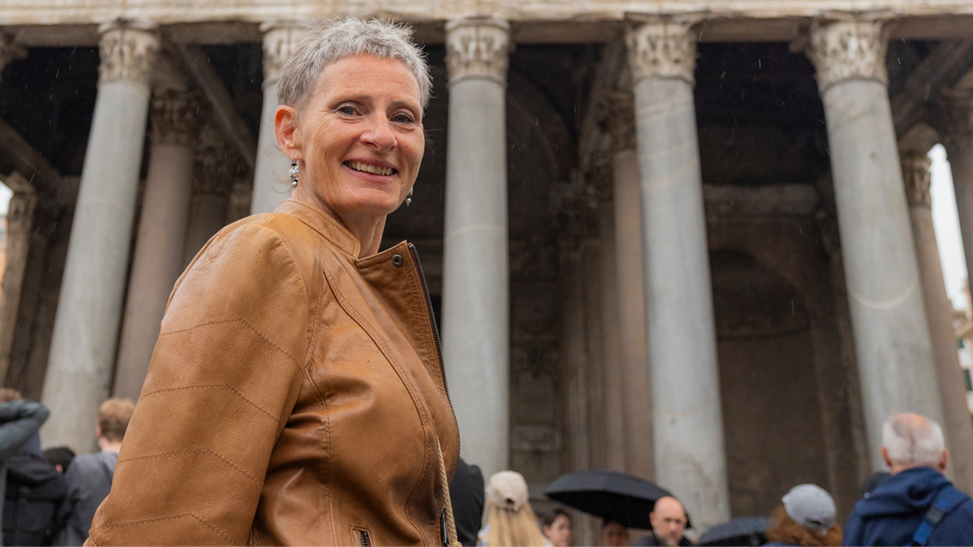A woman with short grey hair smiling in front of the Pantheon in Rome, with a crowd of visitors gathered on the square behind her.