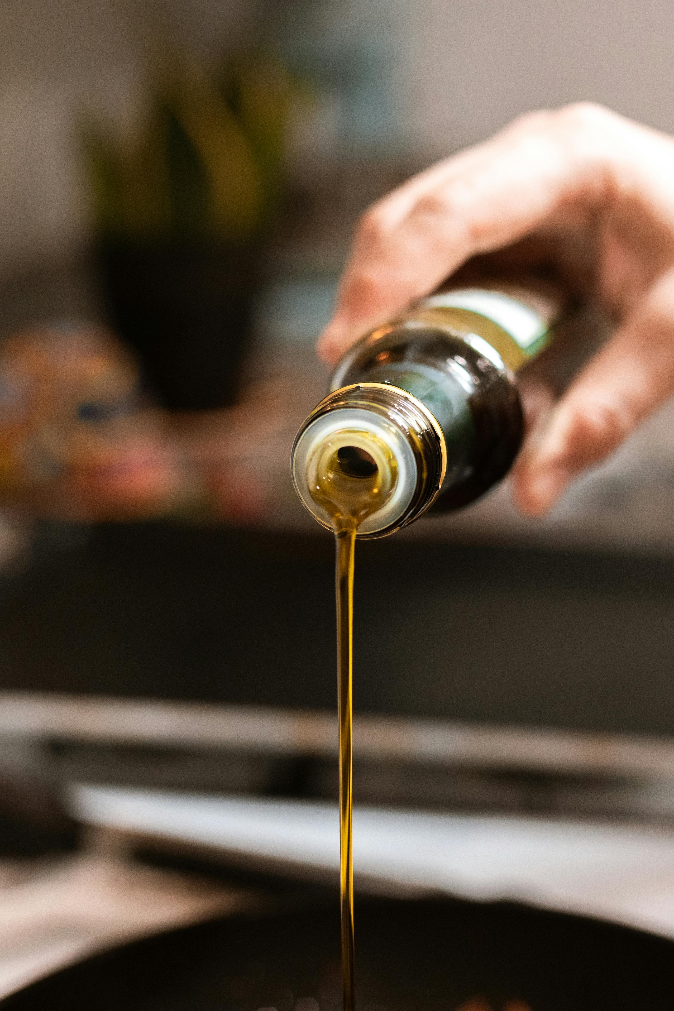 Nahaufnahme einer Hand, die Rizinusöl aus einer Flasche in eine Pfanne gießt, mit unscharfem Küchenhintergrund.