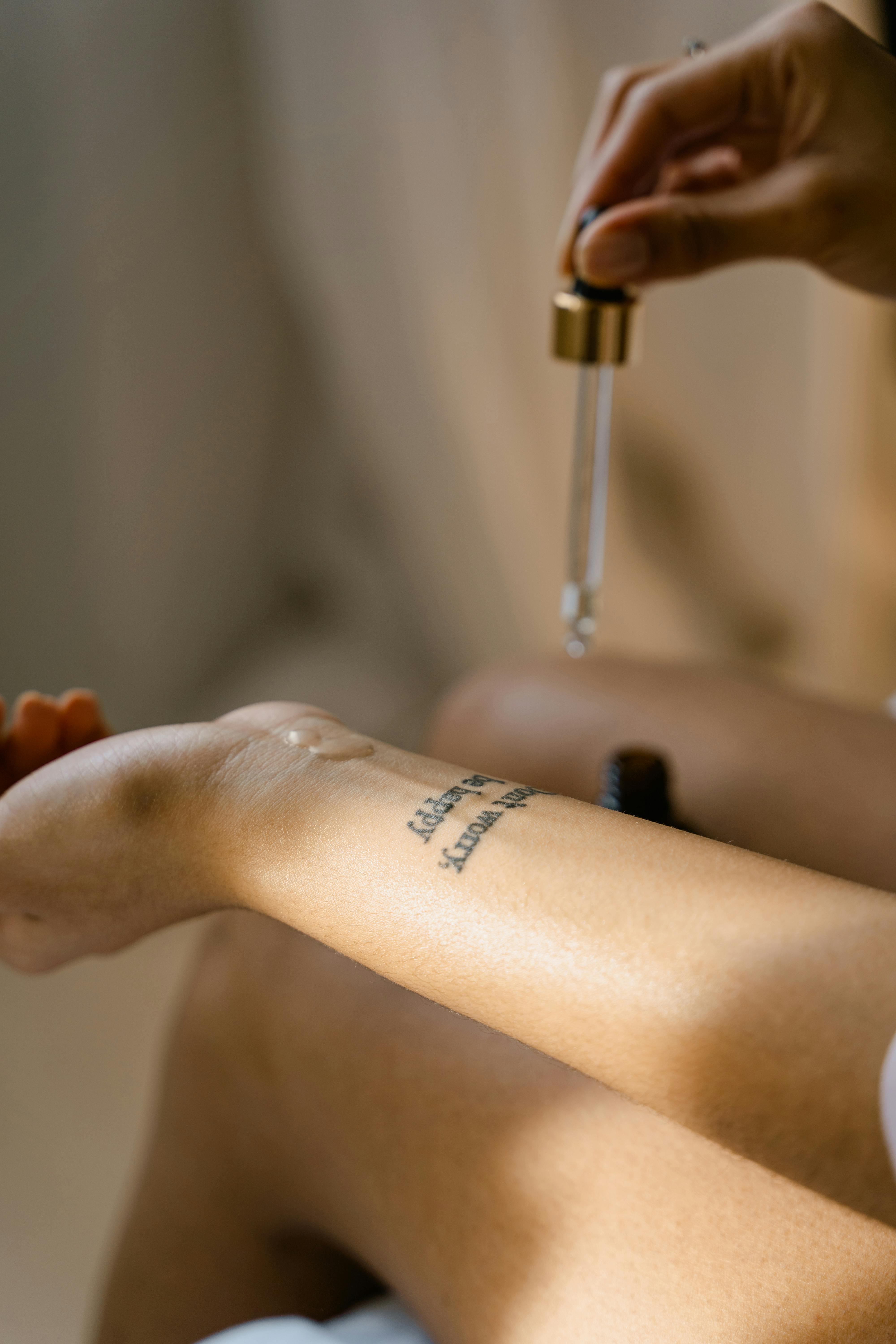 A person applies oil from a dropper onto their forearm, which has a tattoo. The scene is softly lit, creating a calm atmosphere.