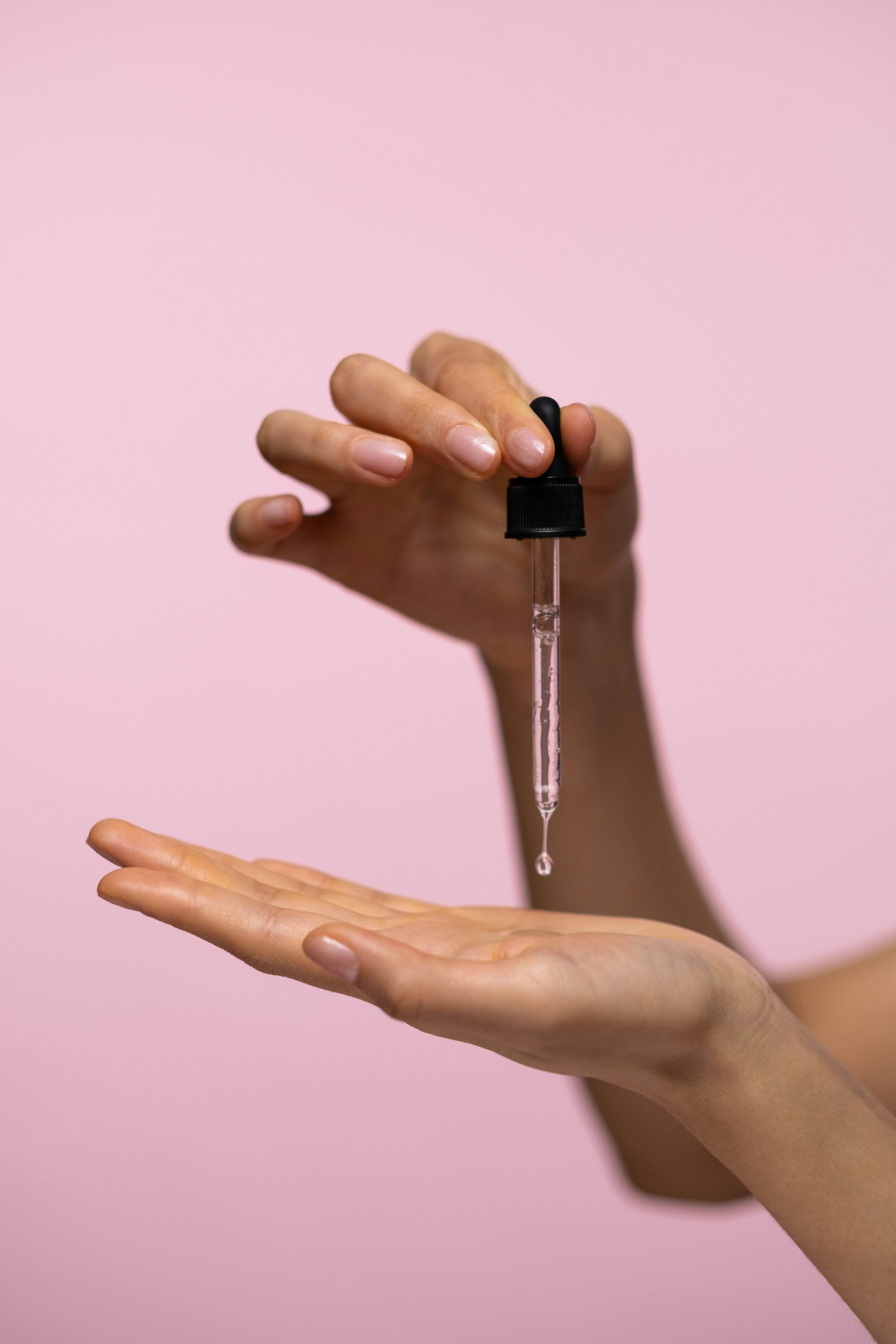 Hände, die eine Pipette halten und Flüssigkeit auf die Handfläche tropfen lassen, vor einem rosa Hintergrund.