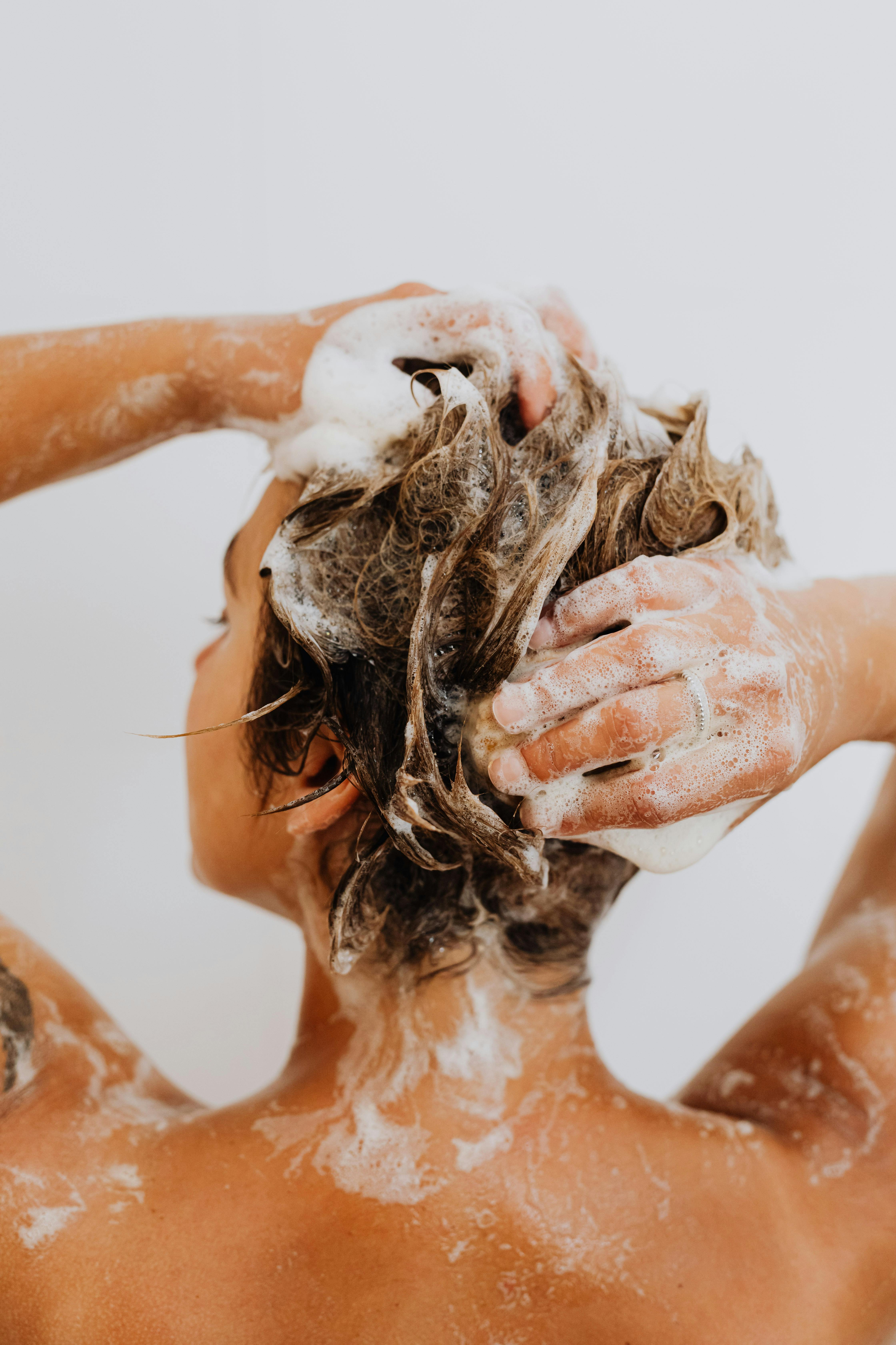 Person, die sich die Haare mit schäumendem Shampoo wäscht, dabei mit den Händen die Kopfhaut massiert und Wassertropfen auf der Haut sichtbar sind.