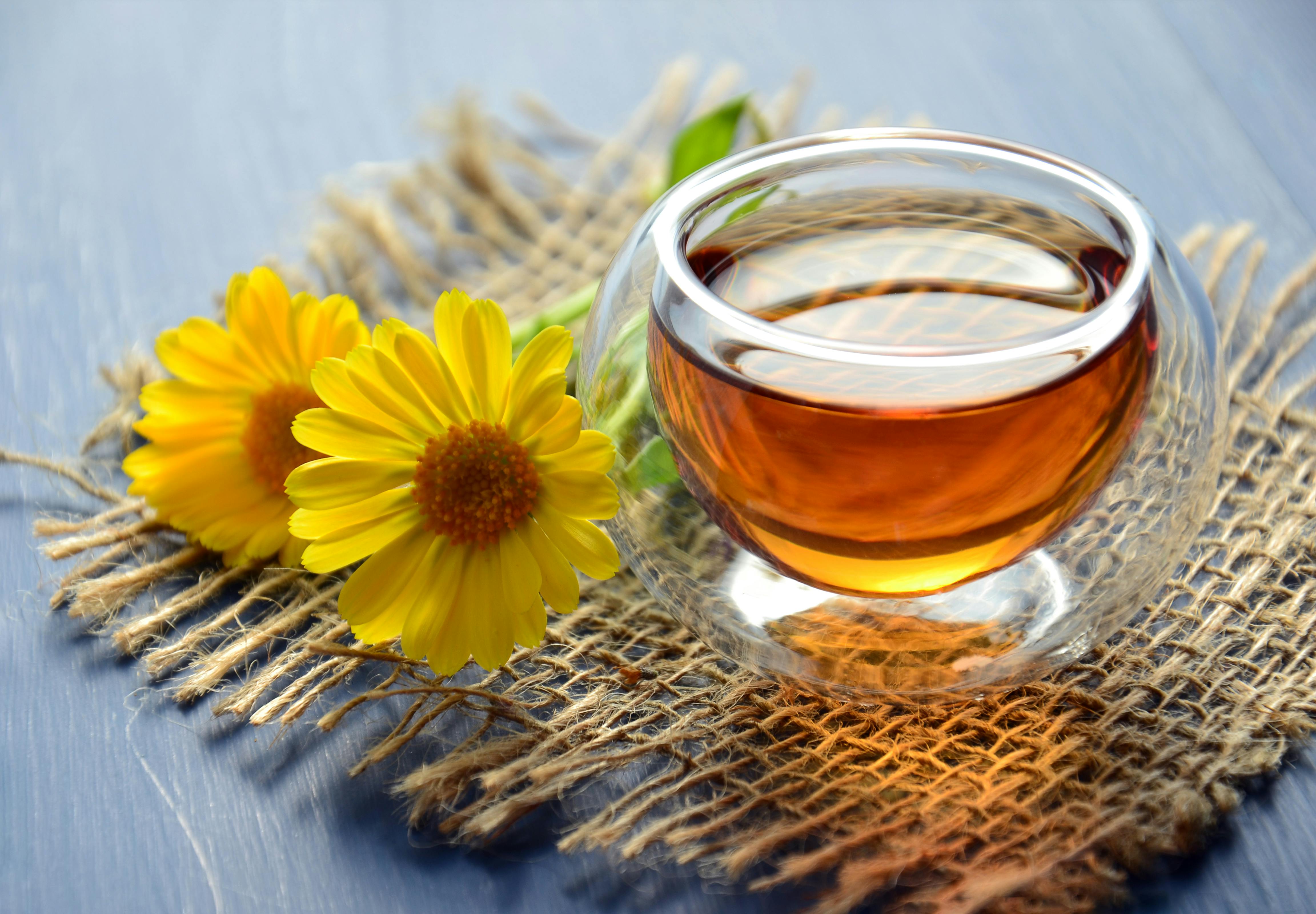 Eine Glas-Tasse Tee auf einer geflochtenen Matte, begleitet von zwei leuchtend gelben Blumen auf einer blauen Holzoberfläche.
