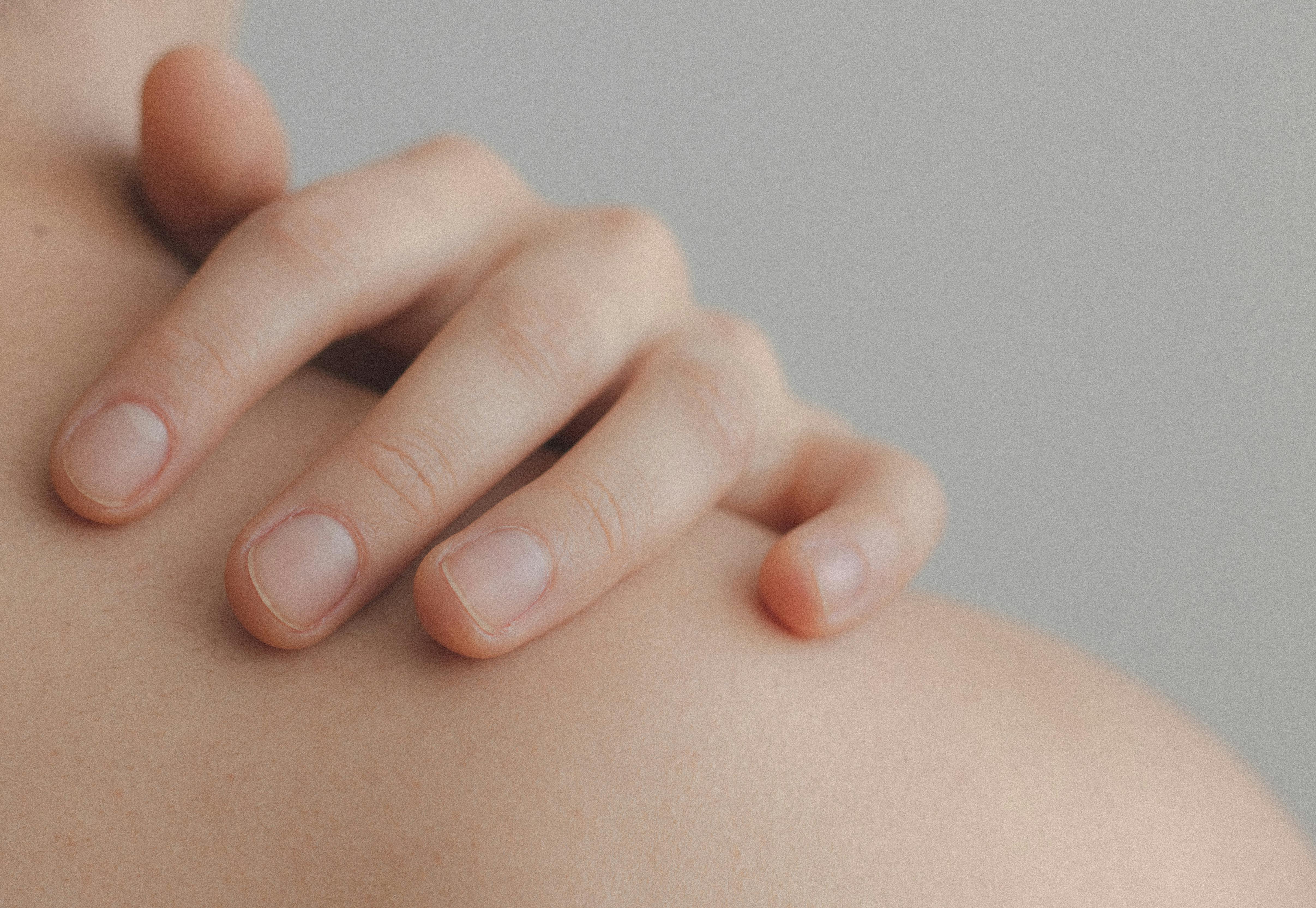 Close-up of a hand gently resting on a bare shoulder against a soft gray background, highlighting smooth skin and natural nails.