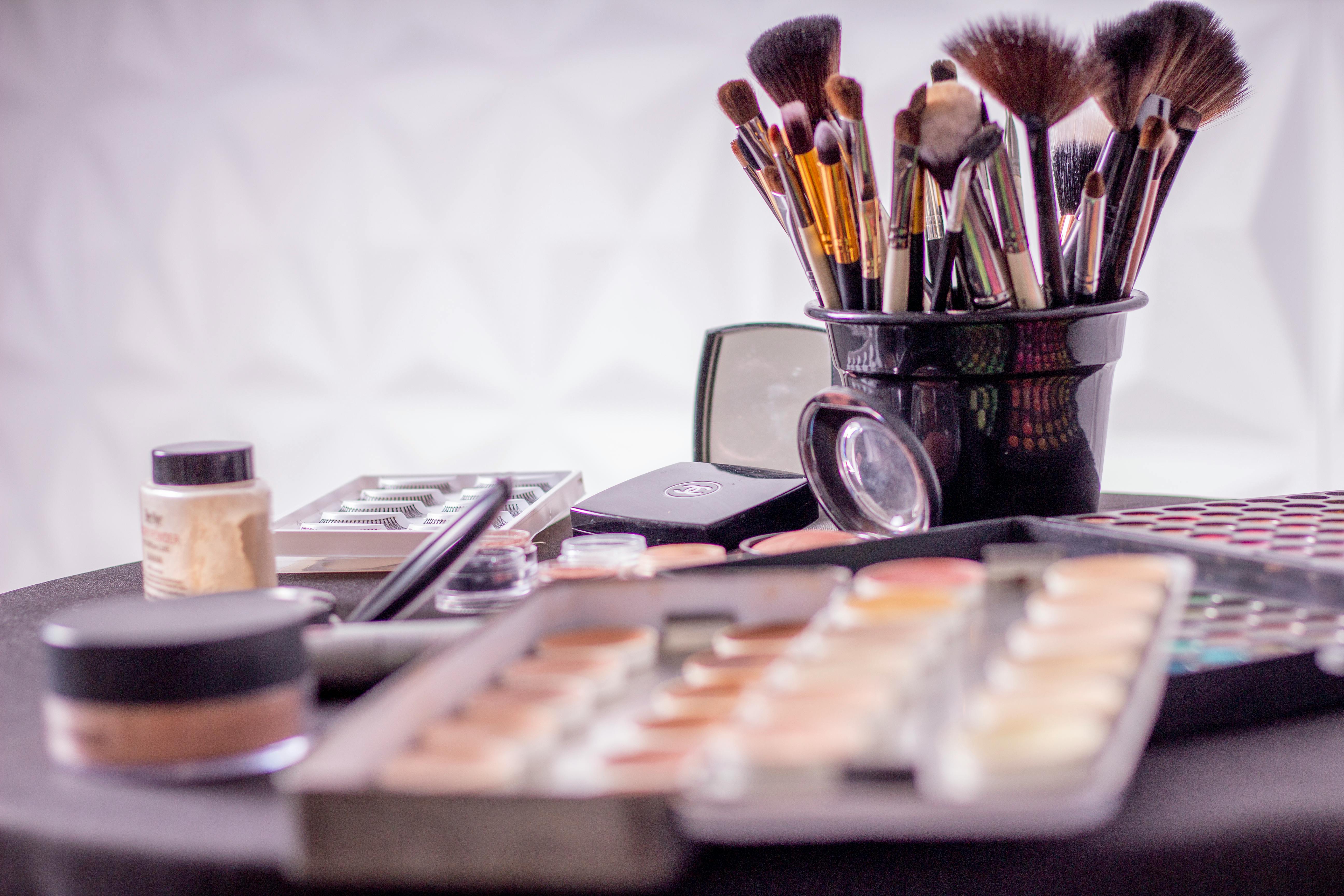 Verschiedene Make-up-Produkte und Pinsel auf einem Tisch, darunter Paletten, Concealer und ein Spiegel, vor einem weichgezeichneten Hintergrund.