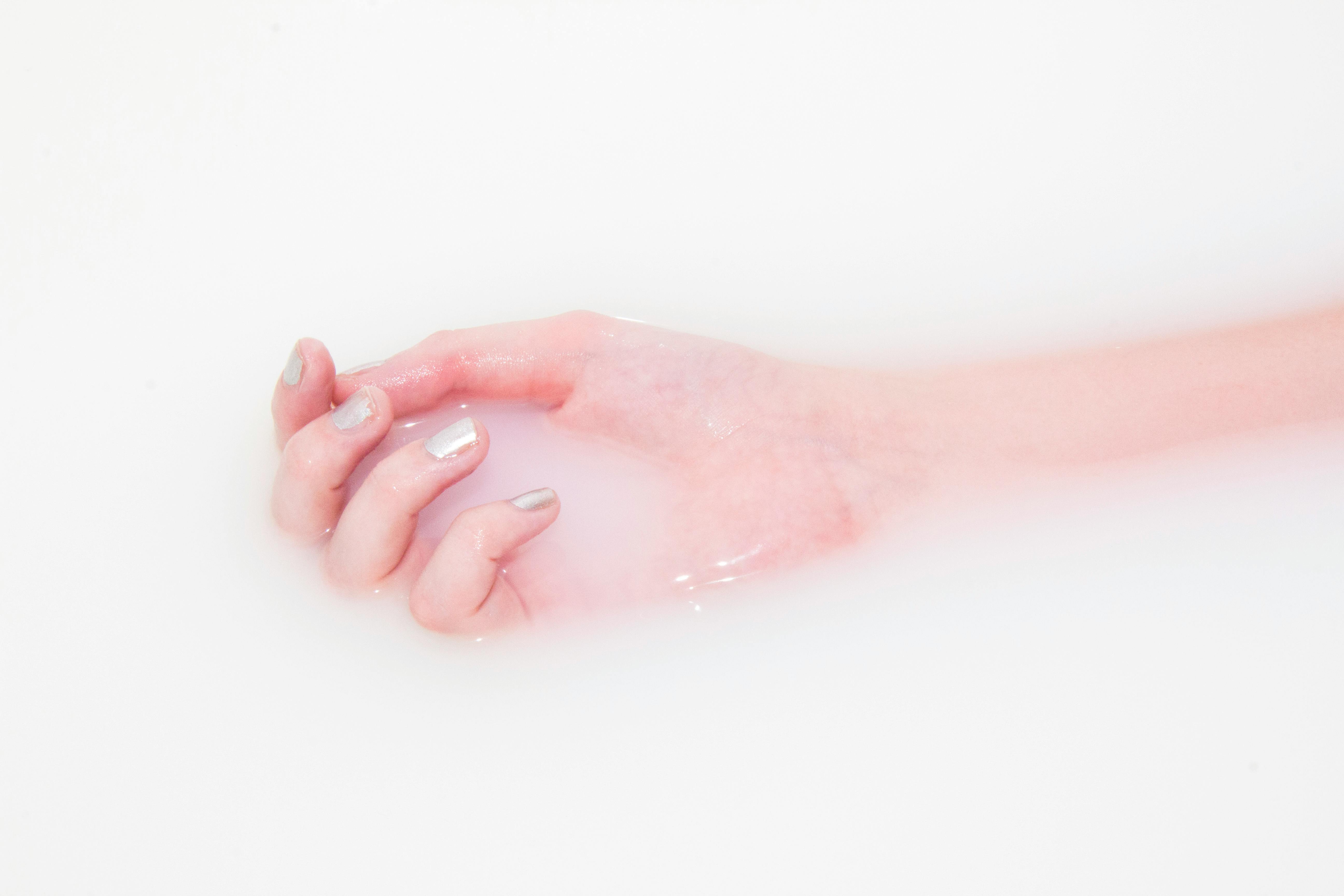 Eine Hand mit blassrosa Nägeln, die teilweise in milchig-weiße Flüssigkeit eingetaucht ist, wodurch ein sanftes, ätherisches Erscheinungsbild entsteht.