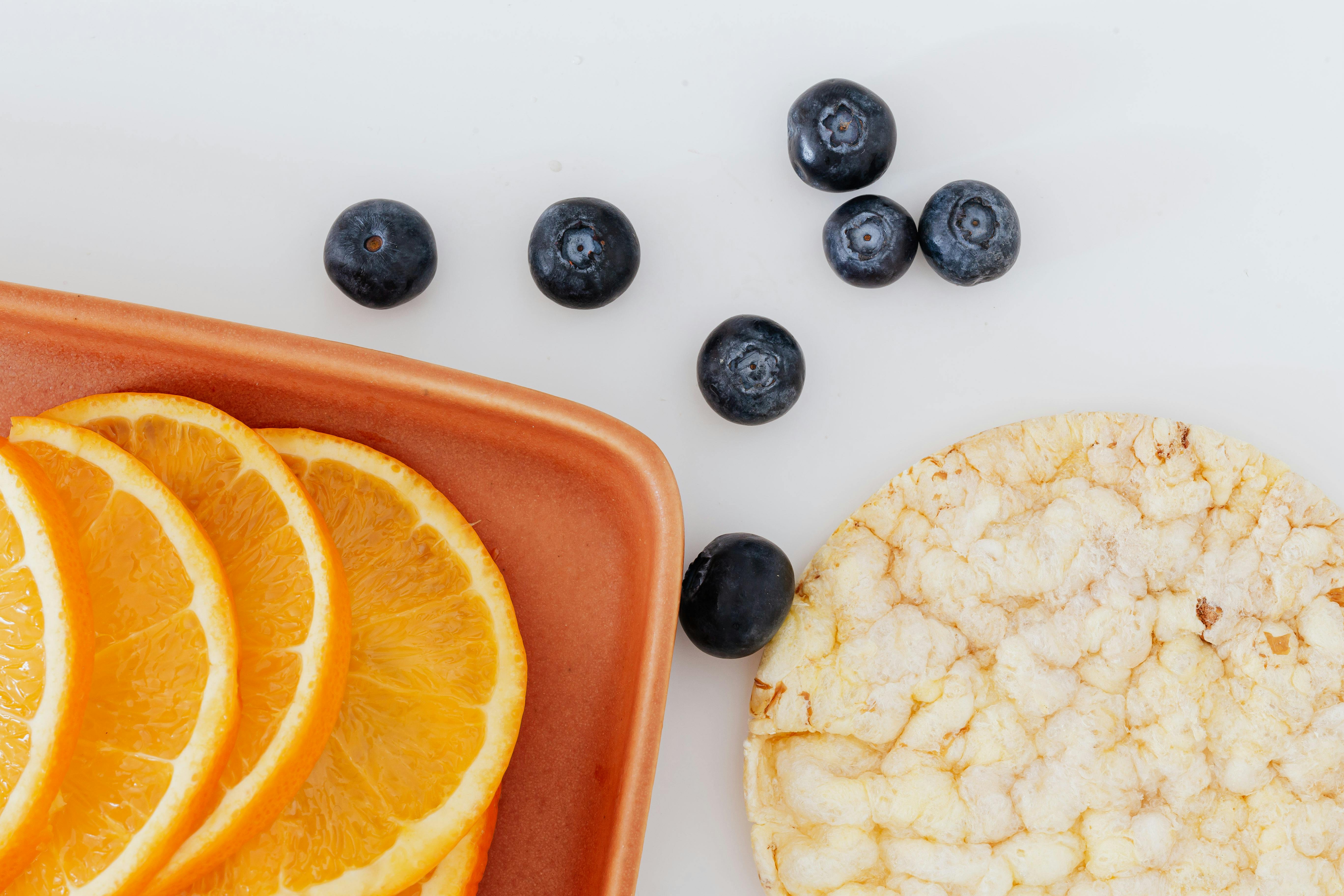 Sliced oranges on a brown plate, blueberries scattered, and a plain rice cake on a white background.