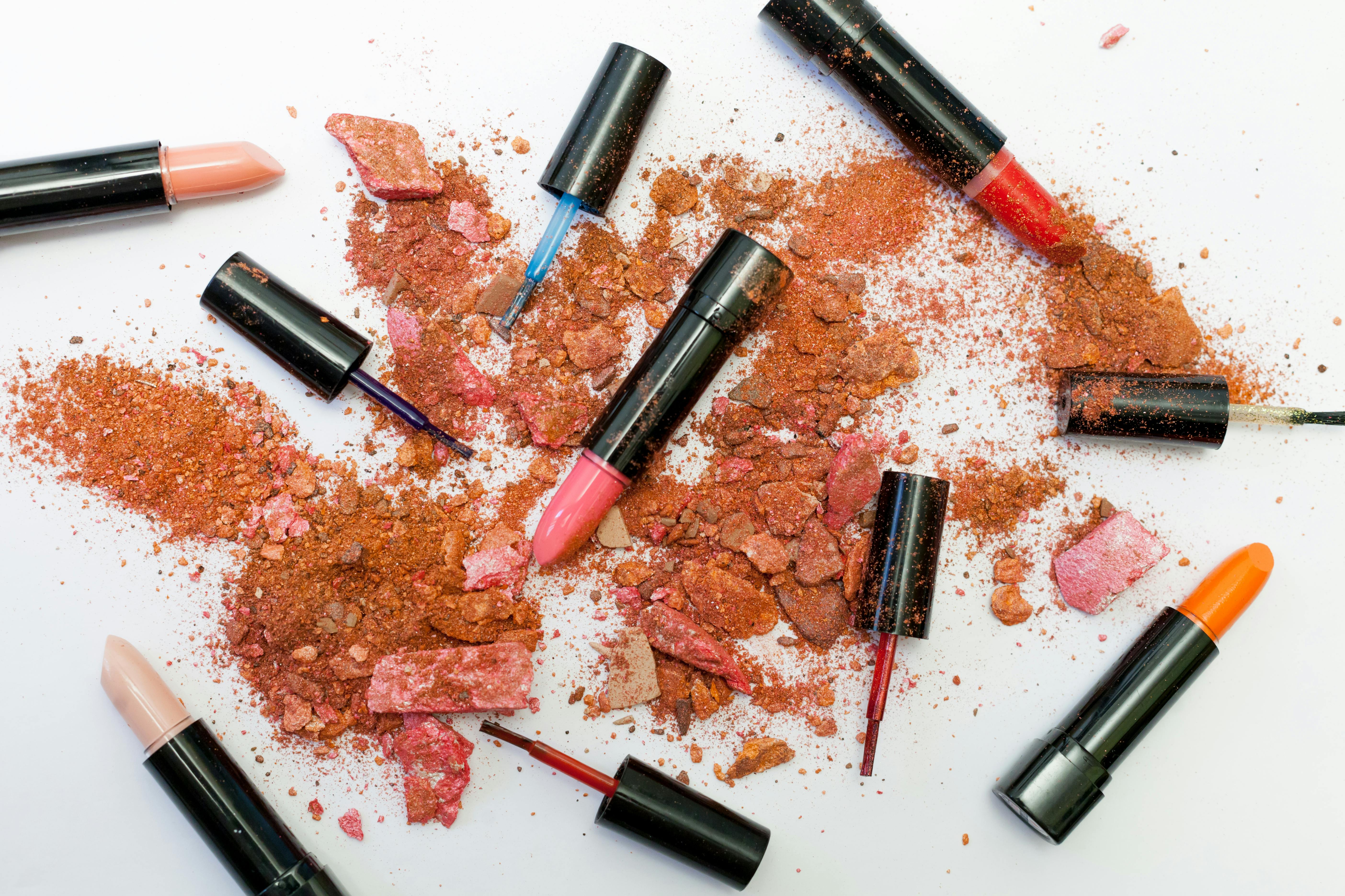 Auf einer weißen Oberfläche verstreute, zerkleinerte Make-up-Puder und verschiedene Lippenstifte, die lebendige Farben und Texturen zur Geltung bringen.