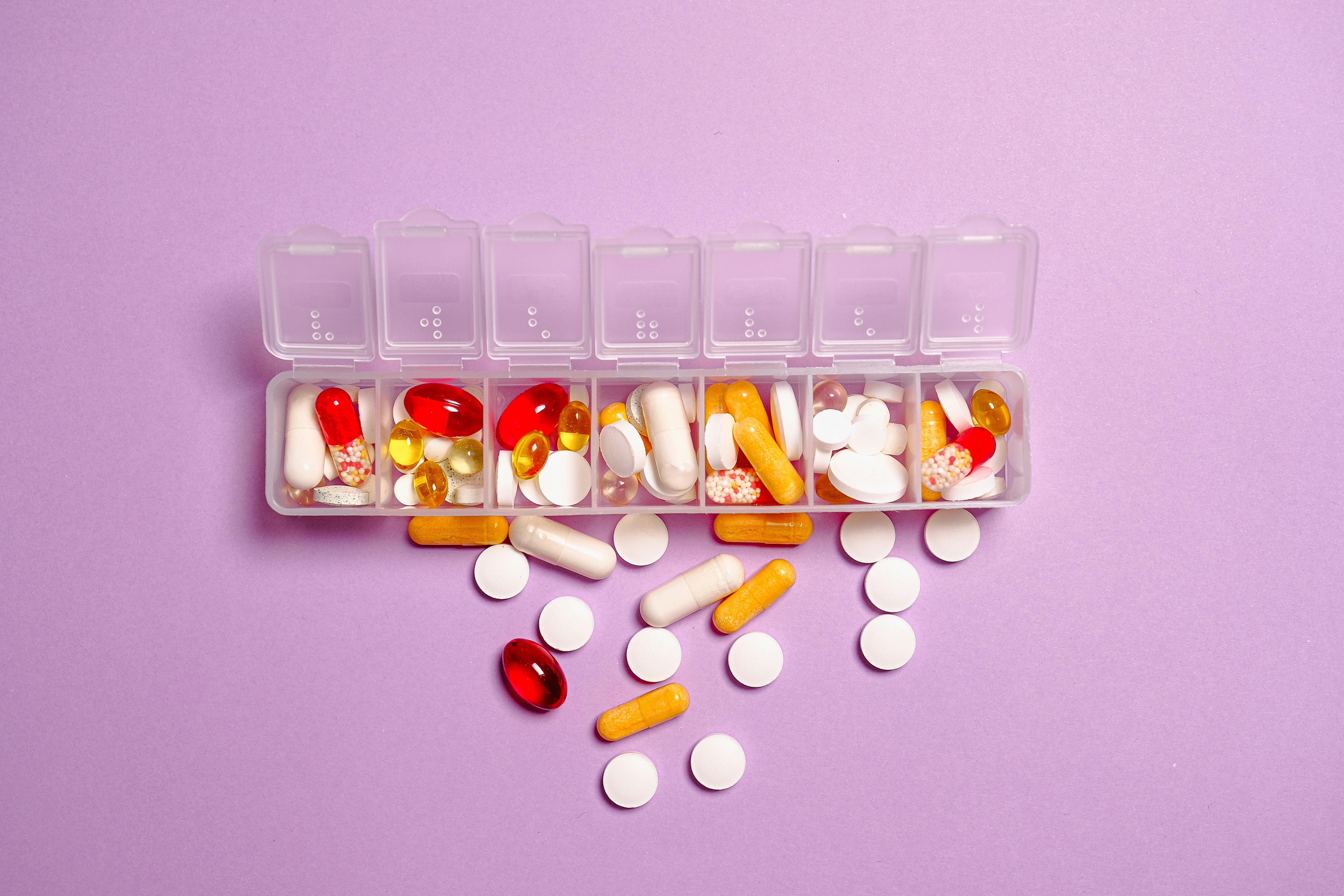 Ein wöchentlicher Pillenspender mit verschiedenen bunten Pillen und Kapseln, die auf einer violetten Oberfläche verstreut sind.