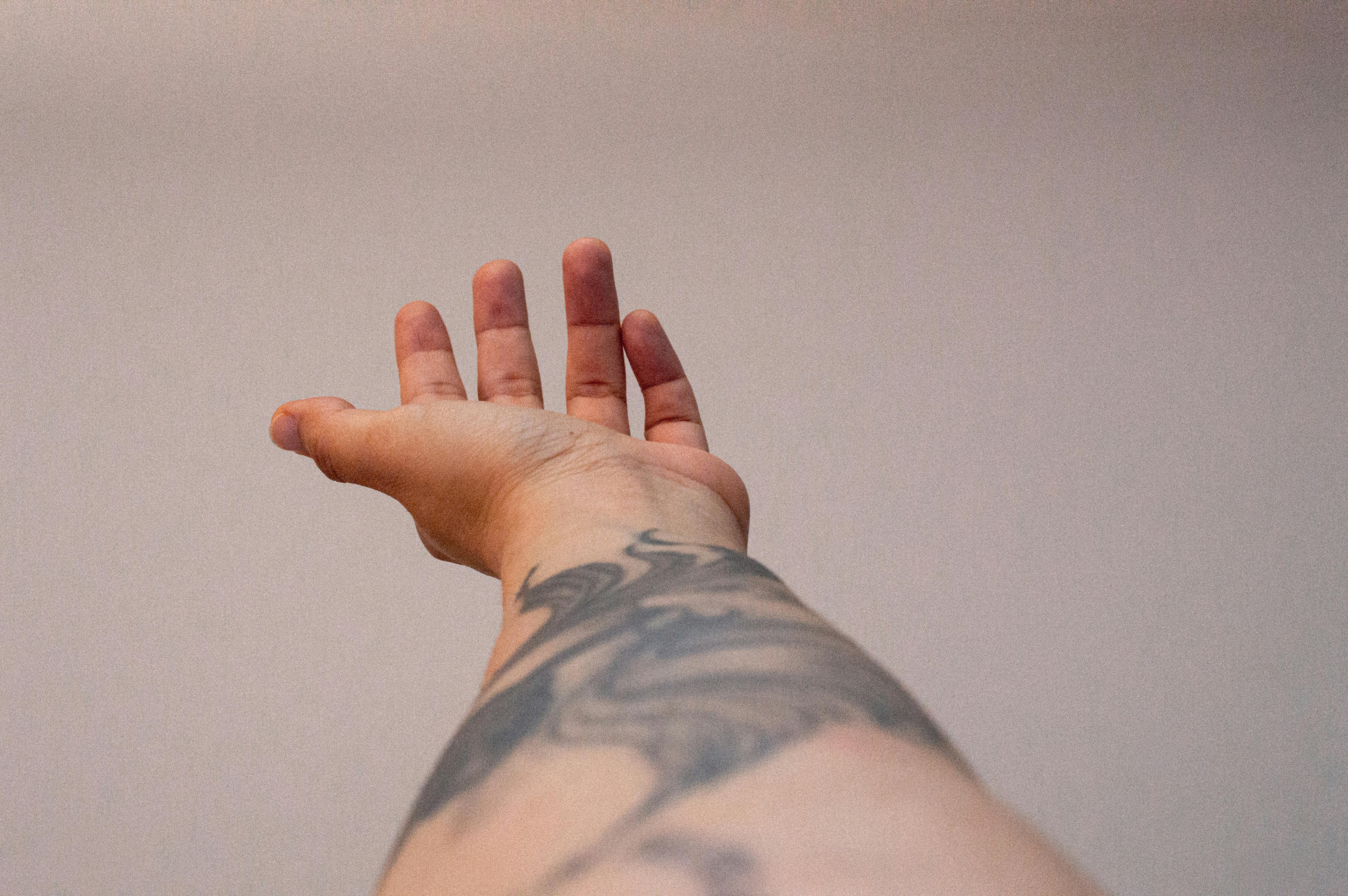 Tätowierter Unterarm, der sich vor einem einfarbigen Hintergrund nach oben streckt, Finger leicht gespreizt.
