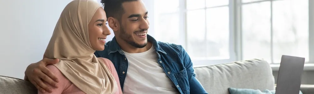 A smiling couple sitting on a sofa, using a laptop together to look at a conveyancing quote from My Home Move Conveyancing in a bright living room. The woman wears a hijab, and the man casually rests his arm around her.