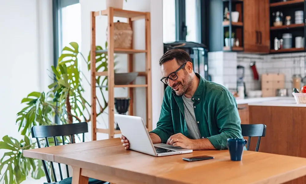 middle aged man smiling and sitting at this kitchen table checking his competitively priced conveyancing quote on his laptop