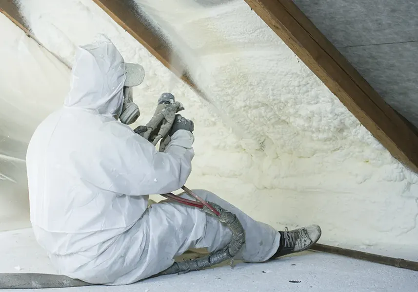 Worker in protective gear sprays foam insulation in an attic, seated on the floor, surrounded by wooden beams and white insulating foam.