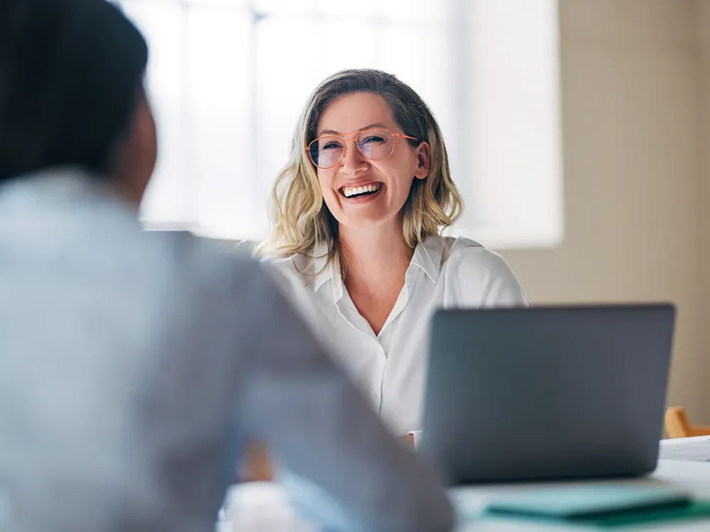 business woman sat at her desk with a laptop in front of her smiling and laughing with the man on the other side of the desk