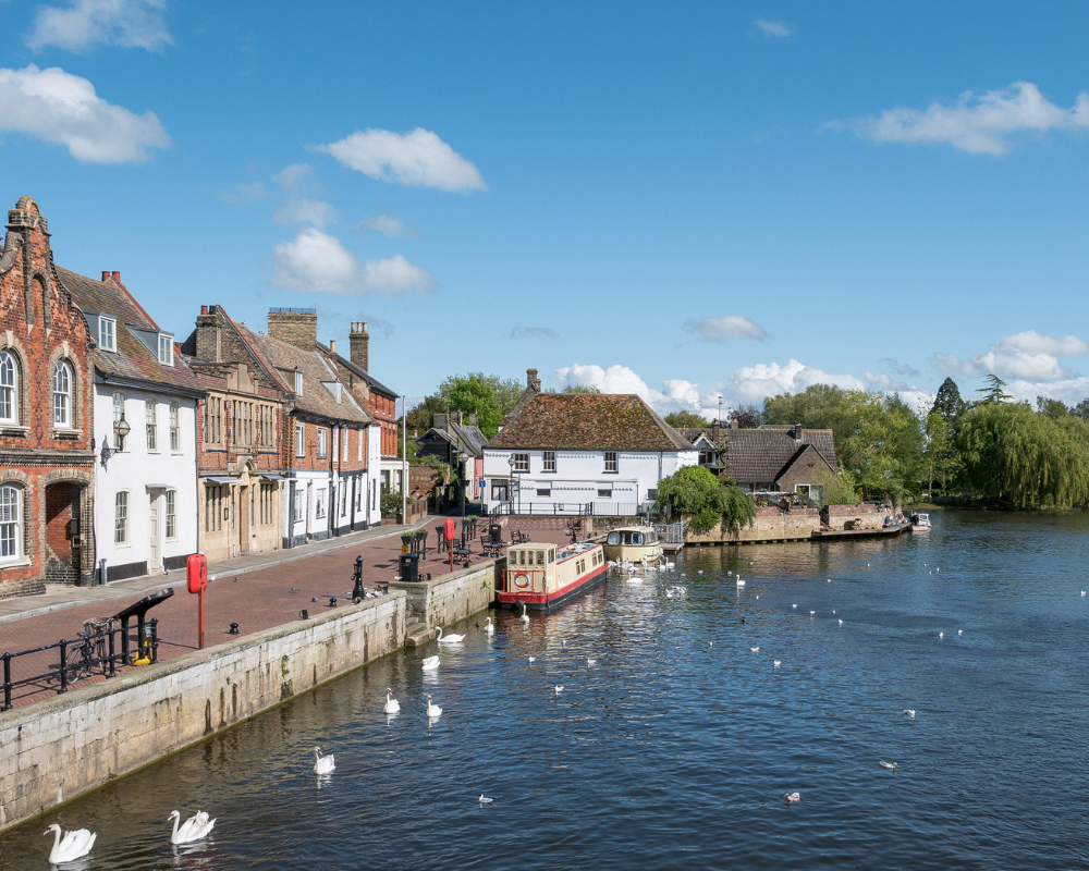 riverside road of old houses in St. Ives in Cambridgeshire. Its a sunny day with a few fluffy clouds in the sky and there are swans on the river