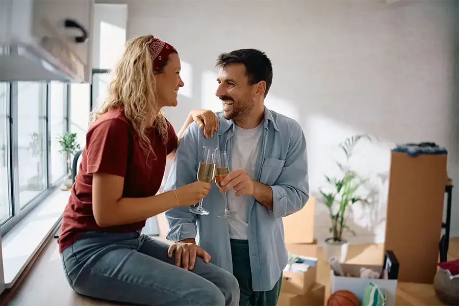 A couple smiles at each other while toasting with wine glasses in a sunlit room filled with moving boxes.