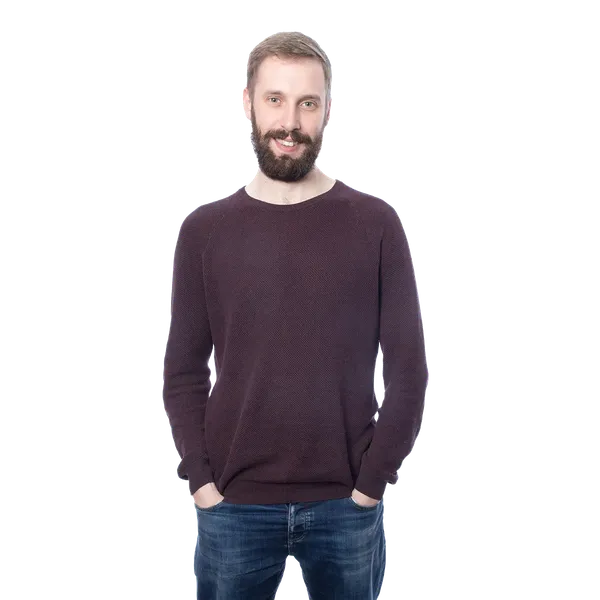 Man with a beard wearing a dark sweater and jeans, standing against a blurred, striped background.