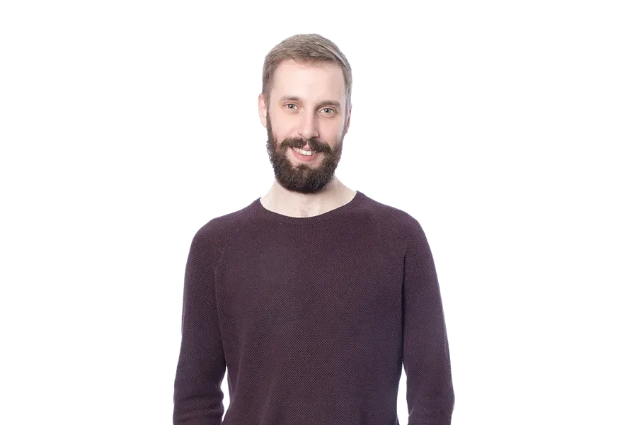 Man with a beard wearing a dark sweater and jeans, standing against a blurred, striped background.